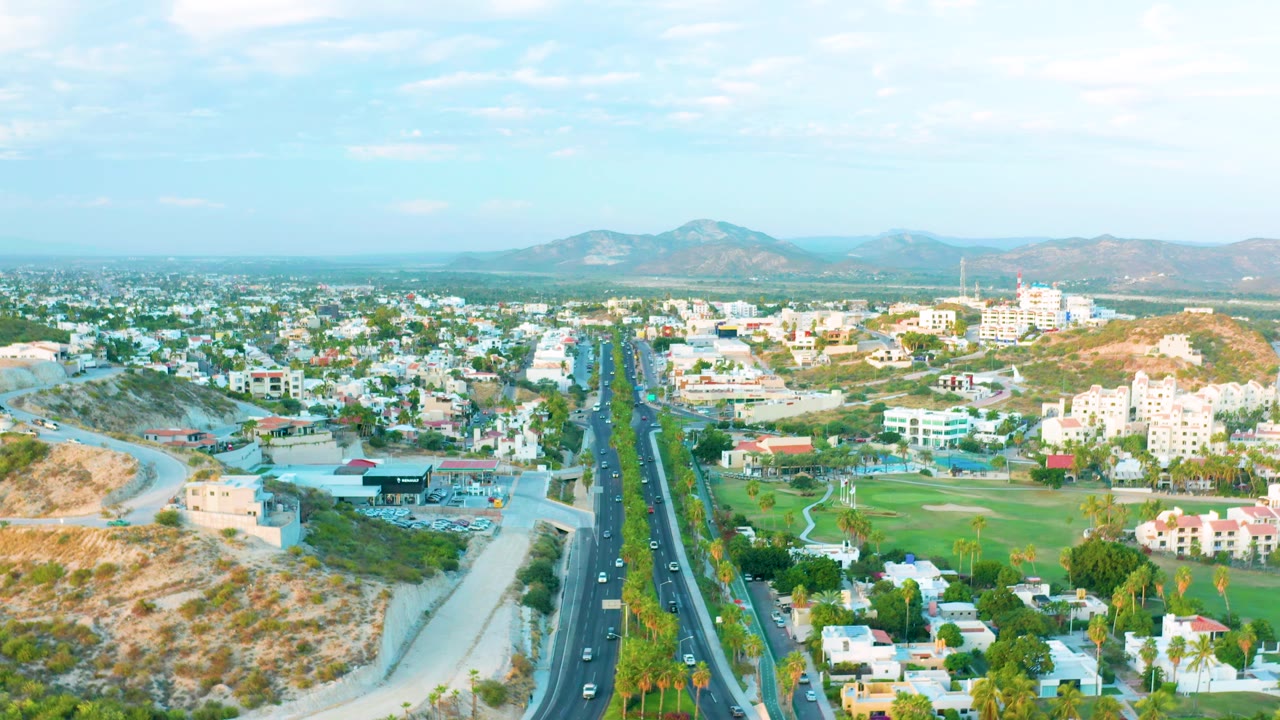 Early aerial view in San Jose Del Cabo with traffic aligned on the center road, white buildings on both sides, tan hills, green landscaping, and no visible people.