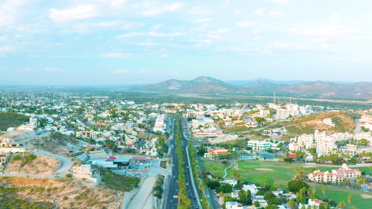 Opening aerial frame over San Jose Del Cabo with a straight divided highway centered between low buildings, dry hills, green areas, distant mountains, scattered clouds, and no visible people.