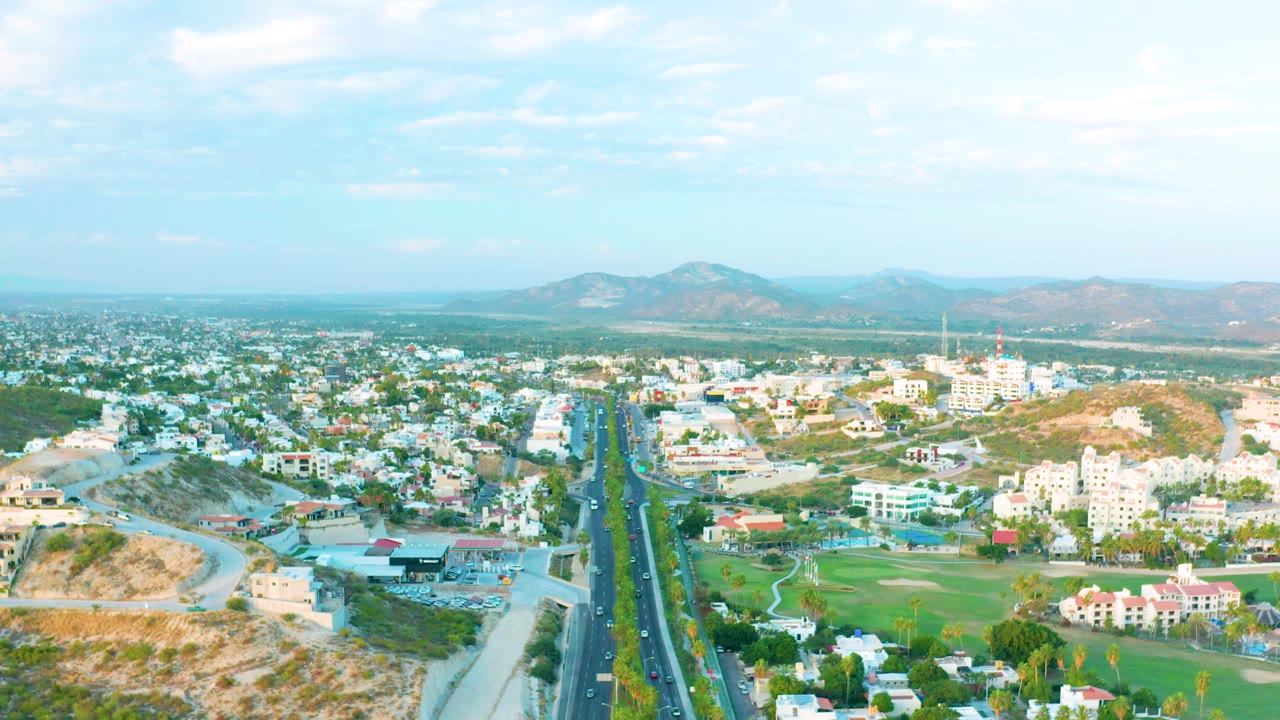 Second aerial frame of San Jose Del Cabo showing the highway slightly farther forward in frame, with white buildings, roadside greenery, dry slopes, mountains on the horizon, and no visible people.