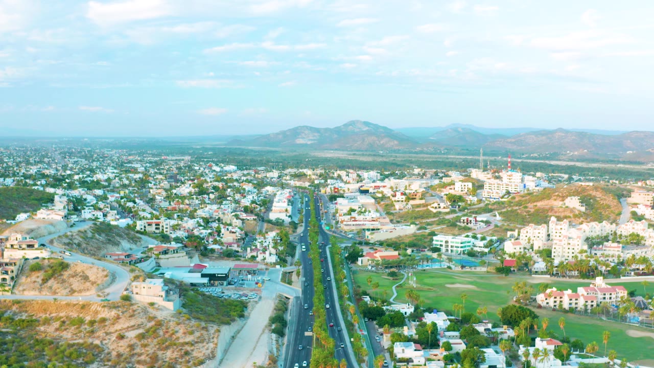 Third aerial frame over San Jose Del Cabo with the divided highway, small moving vehicles, clustered buildings, a green course-like area, dry hillside, distant mountains, and no visible people.