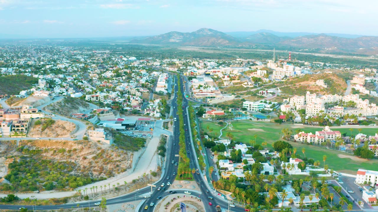 Opening aerial frame over San Jose Del Cabo showing a roundabout at the bottom, a divided road running toward low buildings and distant hills, with no visible people.