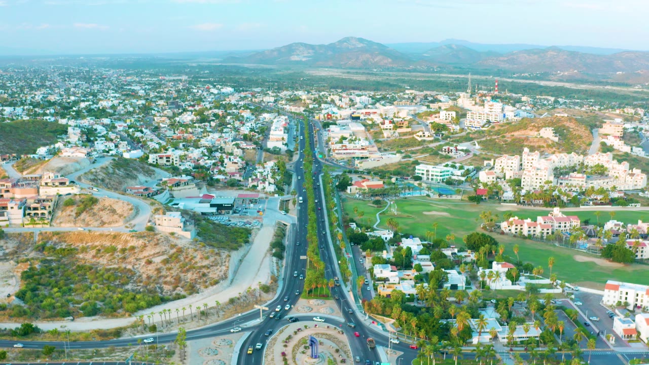 Wide aerial view of San Jose Del Cabo with the central divided road aligned through the frame, a roundabout partly visible below, hillside lots, low buildings, mountains, and no visible people.