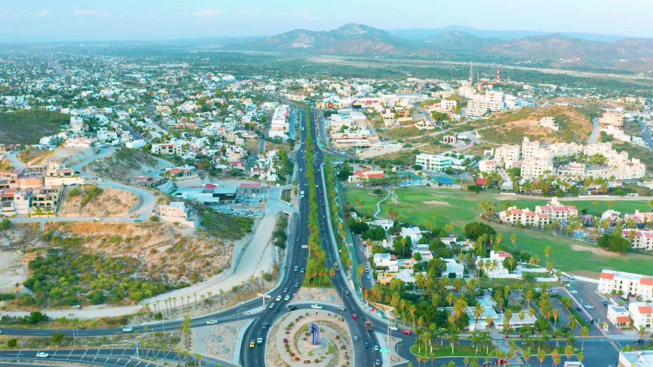Drone view of an urban road, roundabout, homes, green area, and hills.