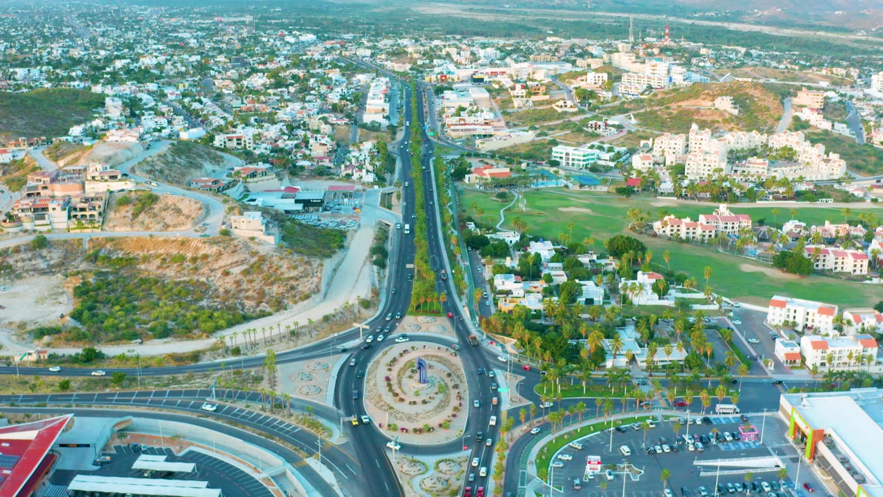 Opening aerial frame over San Jose del Cabo shows a circular roundabout, divided roads, parking areas, green landscaped sections, low-rise buildings, and no visible people.