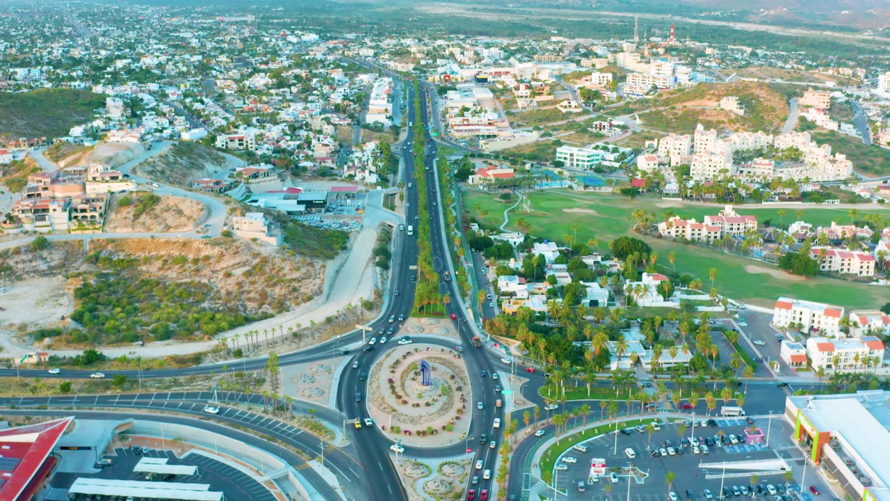 Aerial roundabout with cars, a parking lot, and buildings beyond the road.