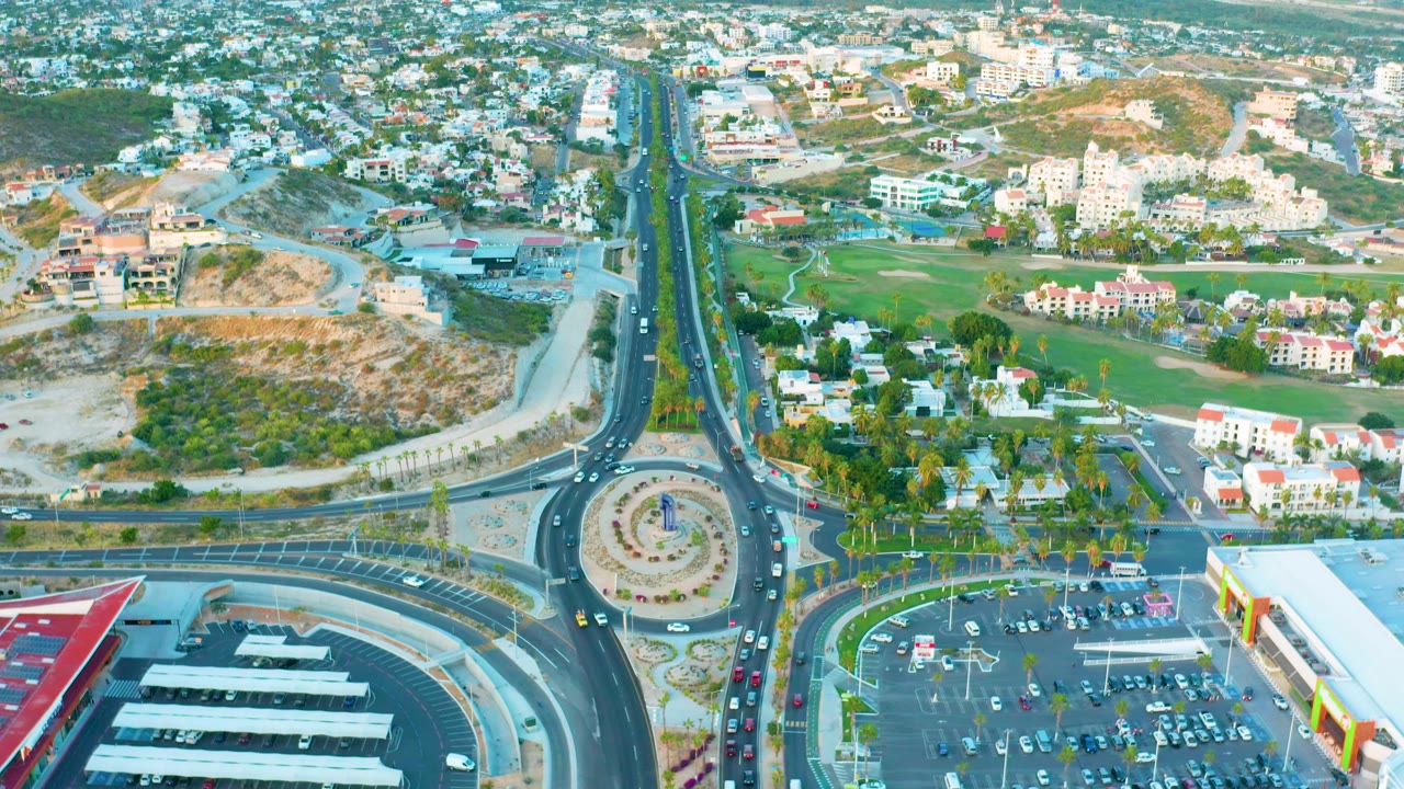 Aerial time-lapse frame of a roundabout beside a full parking lot and low buildings.