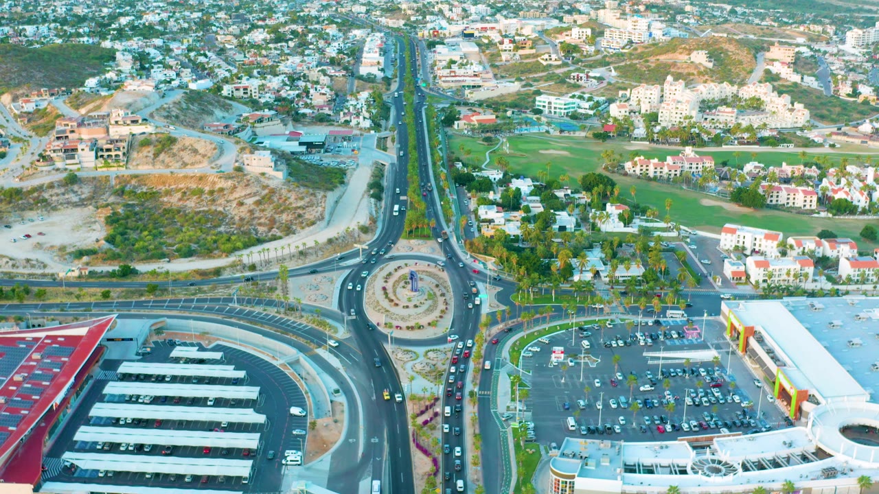 High aerial view in San Jose Del Cabo with vehicles entering and exiting the roundabout, a large parking lot at lower right, covered parking rows at lower left, and no visible people.