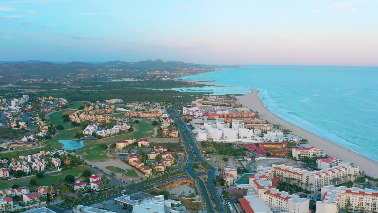 Aerial view in San Jose Del Cabo with clustered resort buildings in the lower frame, a straight road through the center, sandy beach and ocean to the right, hills beyond, and no visible people.