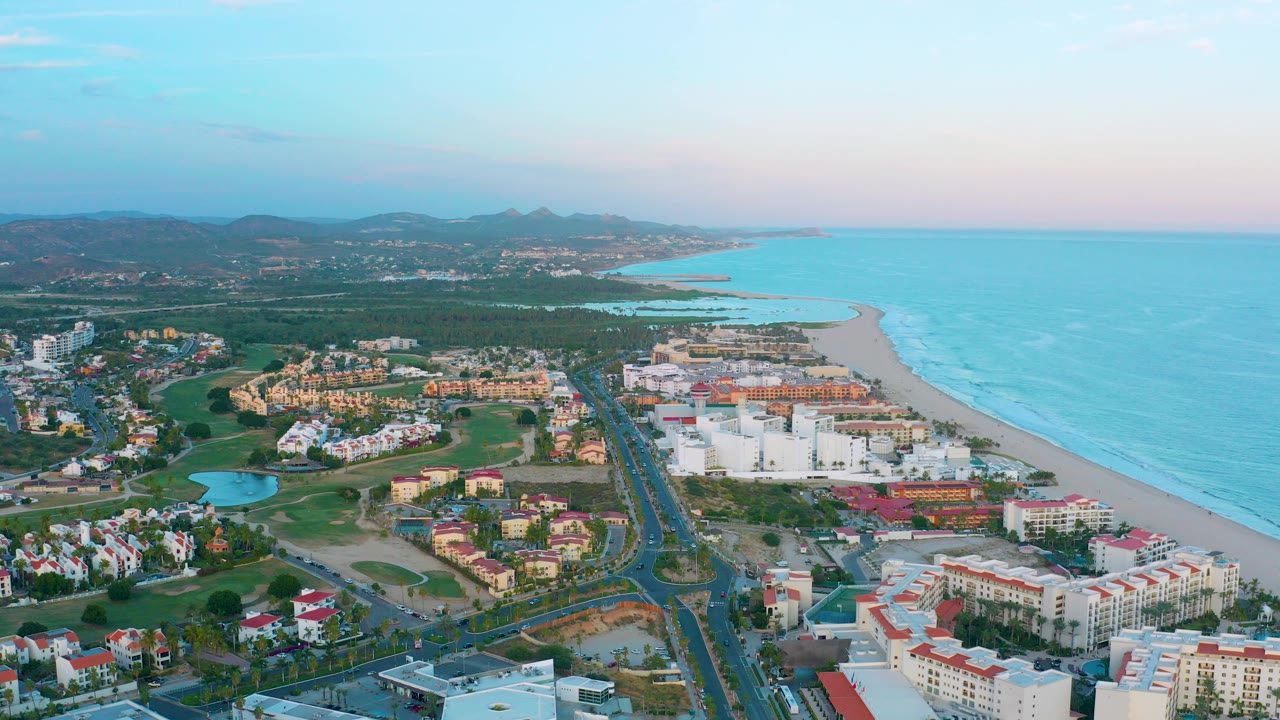 Aerial view over San Jose Del Cabo with resort buildings beside a coastal road, green grounds and a small pond to the left, sandy beach and blue ocean to the right, and no visible people.