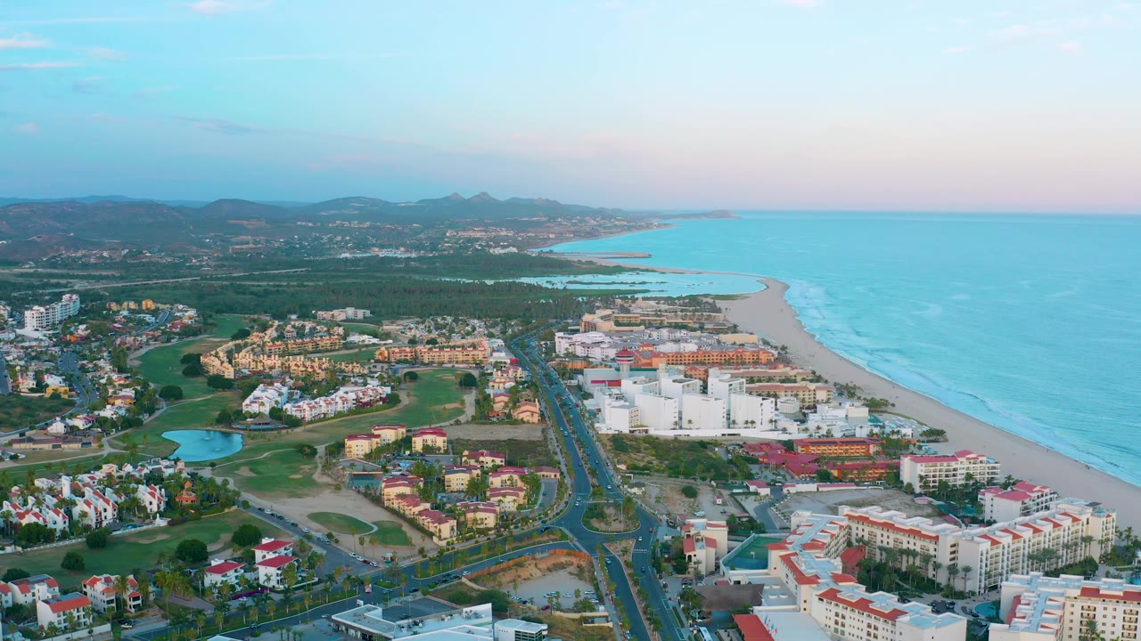 Aerial coastline view with resort blocks, road, beach, surf, and hills