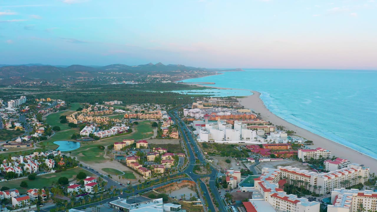 San Jose del Cabo aerial frame showing white resort blocks, a long sandy shoreline, ocean wave bands, a central road, green resort grounds, hills on the horizon, and no visible people.