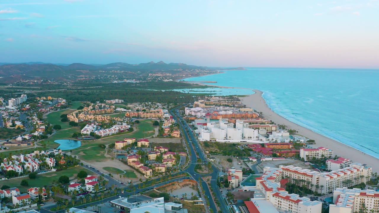 Aerial view over coastal resort area with ocean, beach, road, buildings, and green grounds.