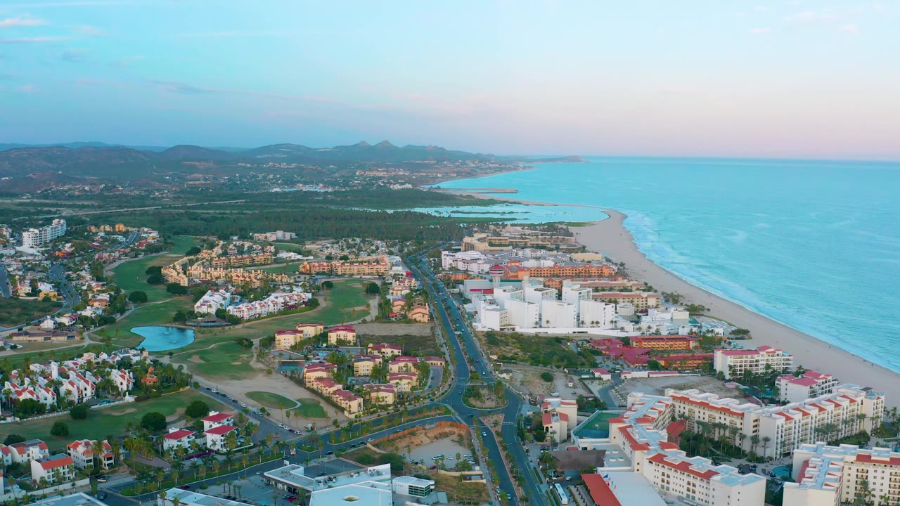 Aerial frame over San Jose del Cabo showing resort-like beachfront buildings, a long road corridor, green open areas, the shoreline, blue water, distant hills, and no visible people.