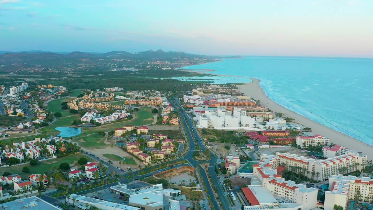 High aerial view of San Jose Del Cabo resort buildings, roads, green grounds, sandy beach, blue ocean, distant hills, and no visible people at the opening moment.