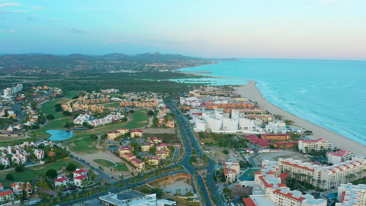 High aerial frame over San Jose Del Cabo resort buildings, a main road, green fairways, sandy beach, turquoise ocean, distant hills, and no visible people.