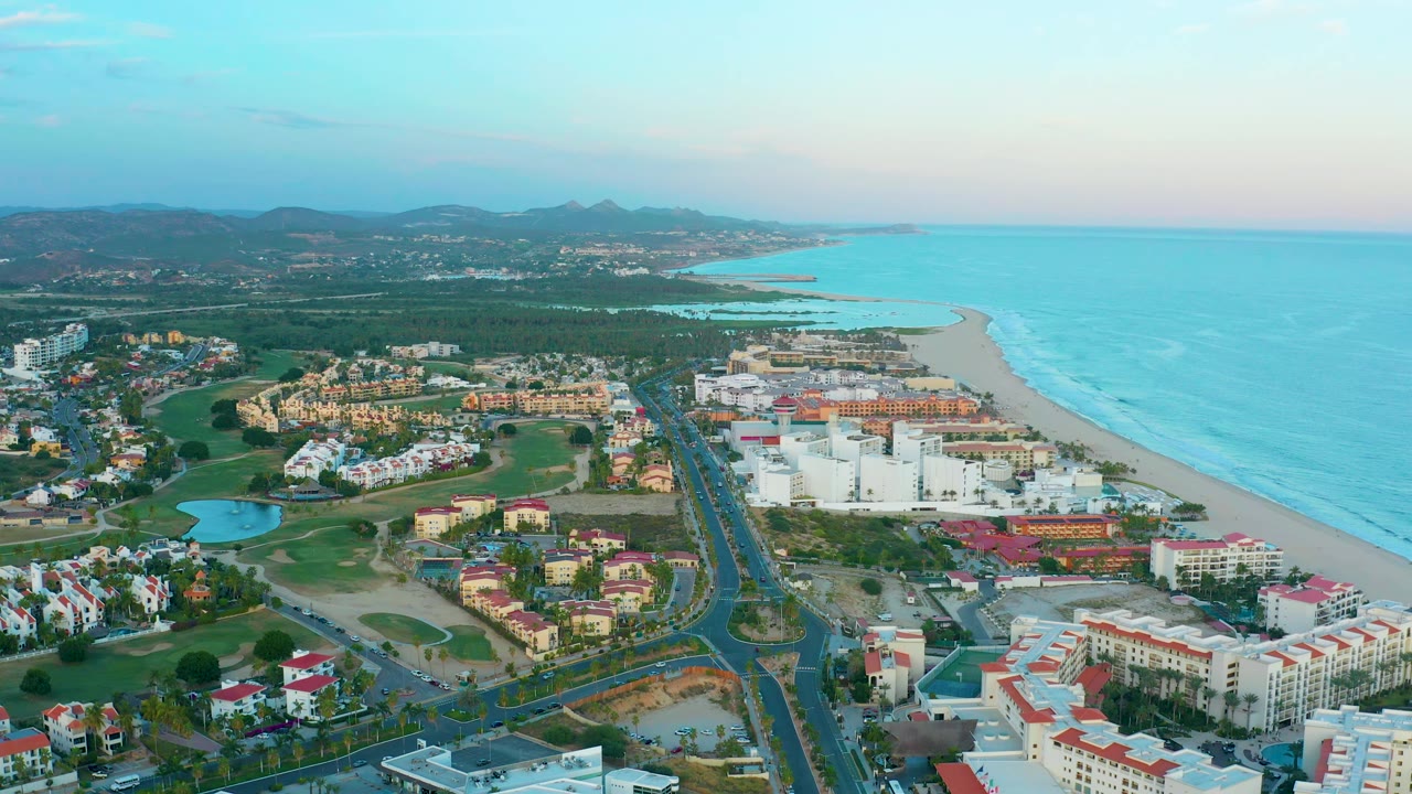 Aerial view in San Jose Del Cabo with beachfront resort blocks, red-roofed buildings, a divided road, golf-course greenery, ocean surf lines, and no visible people.