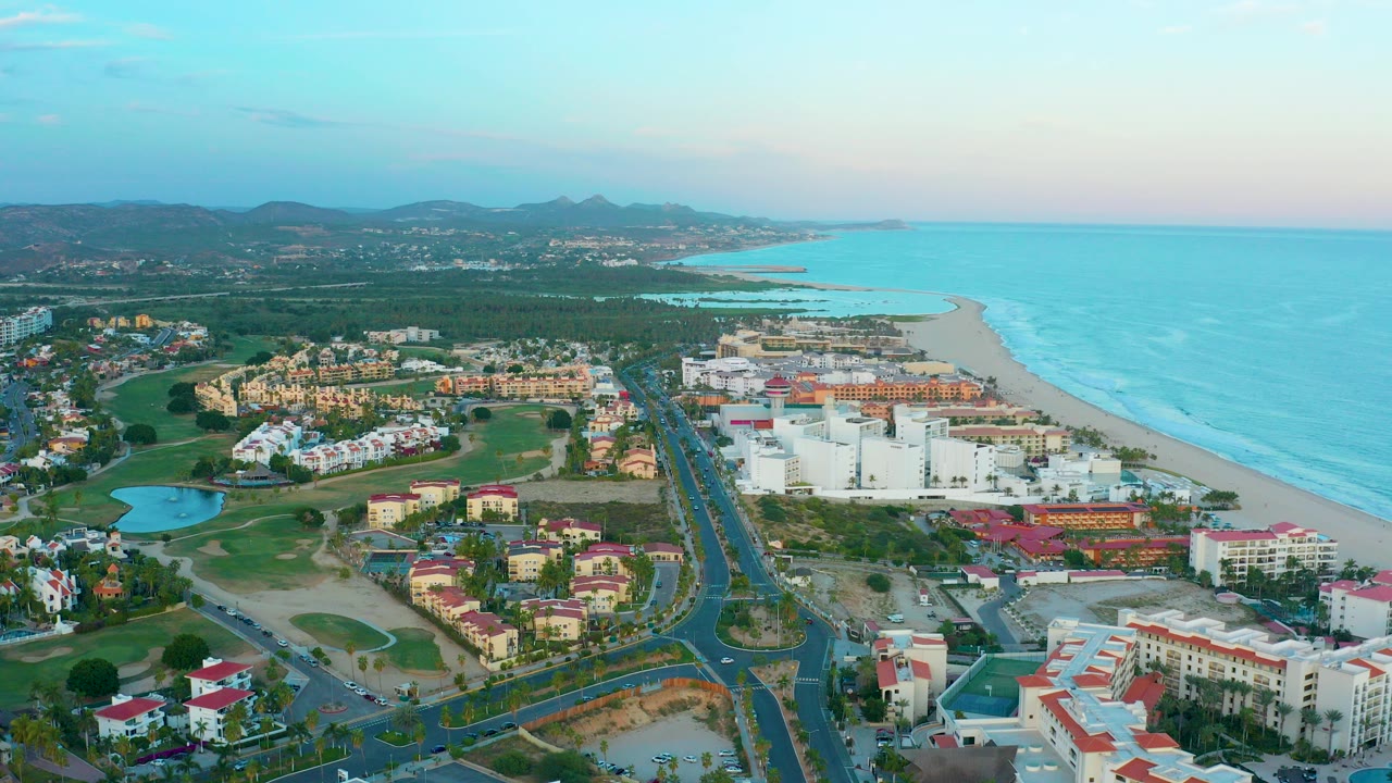 Opening aerial frame over San Jose Del Cabo with resort buildings, green spaces, a road running toward the beach, turquoise ocean to the right, distant mountains, and no visible people.