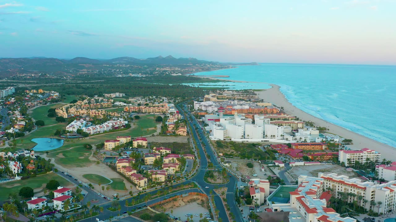 Aerial view in San Jose Del Cabo with the central road and resort buildings slightly more centered, a sandy beach and ocean on the right, hills in the distance, and no visible people.