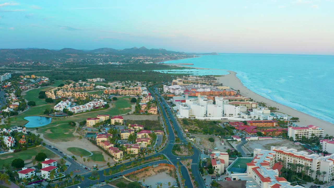Wide San Jose Del Cabo aerial with a small blue water feature at left, clusters of white and tan buildings, the beach curving beside the ocean, distant mountains, and no visible people.