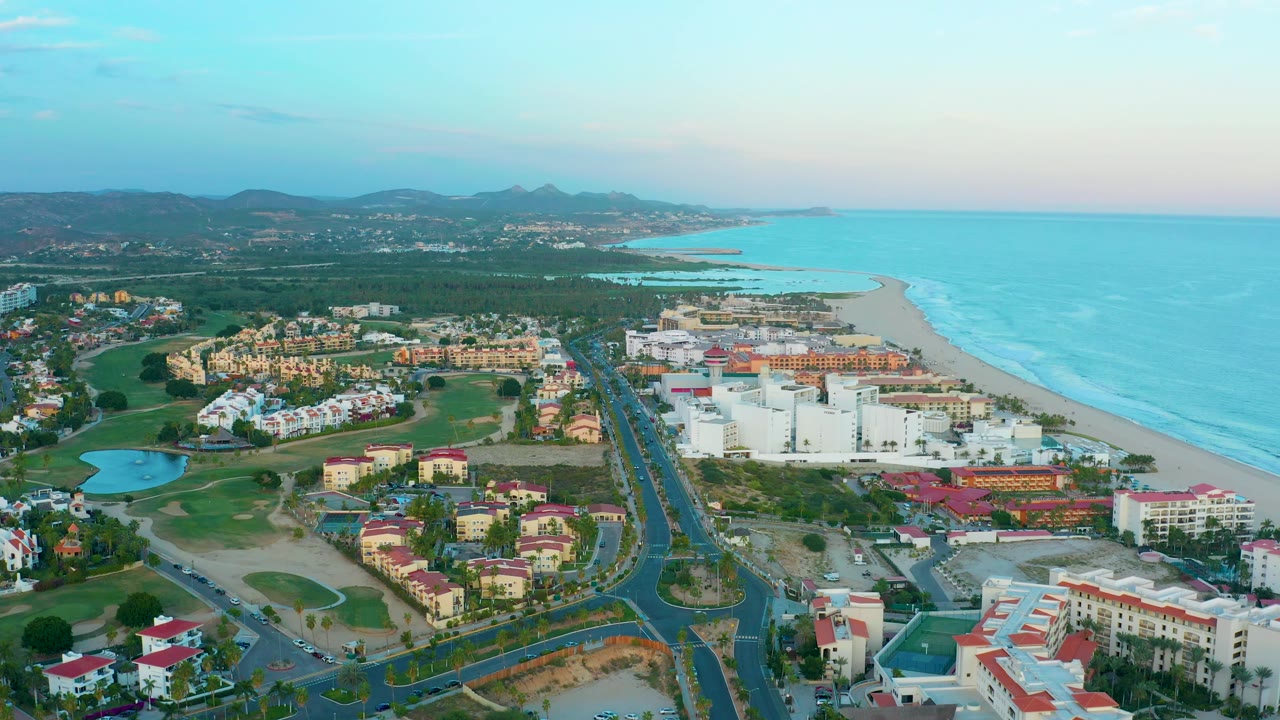 Aerial coastline with resort buildings, road junctions, beach, ocean, and hills.