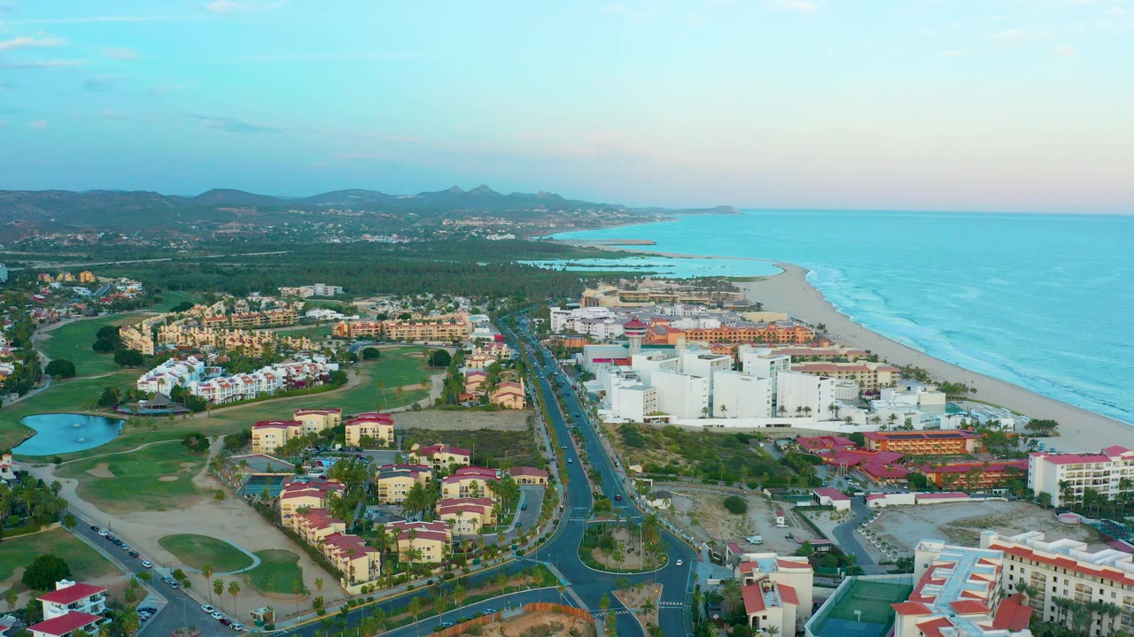 Wide aerial frame over San Jose Del Cabo with golf course greens and red-roofed buildings in front, white resort blocks near the beach, turquoise ocean to the right, distant mountains, and no visible people.