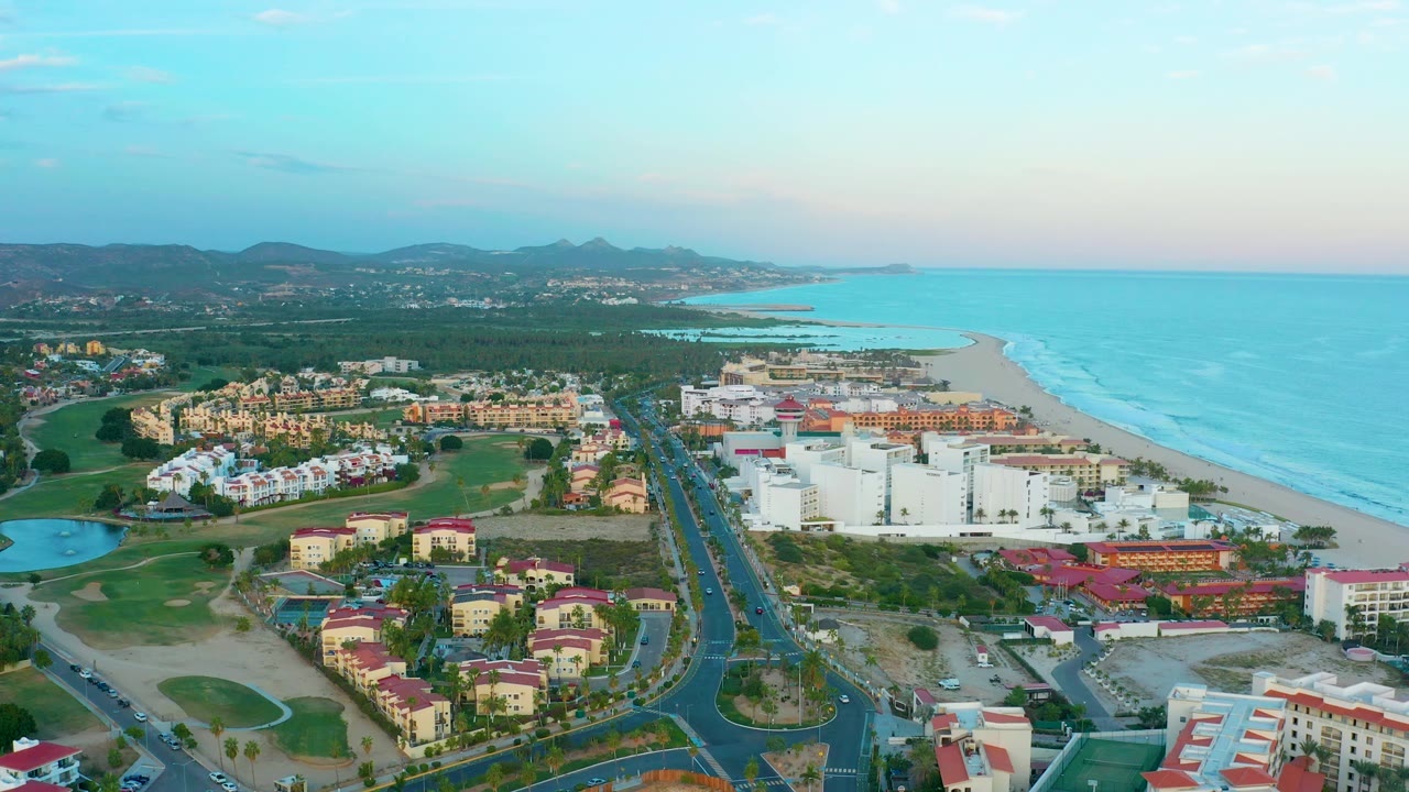 Drone view of resort buildings, divided road, golf course, beach, ocean, and distant mountains.