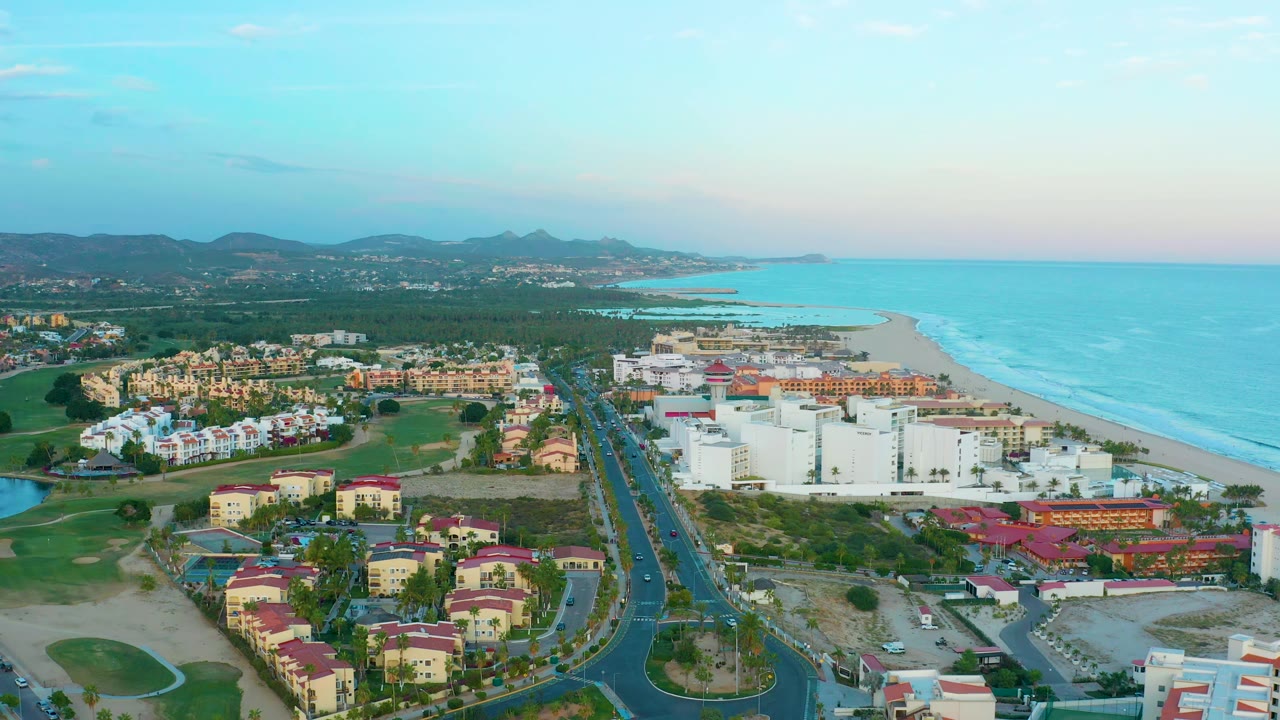 One second in, the San Jose Del Cabo aerial view holds on resort blocks beside the beach, small cars on the central road, turquoise ocean, inland hills, and no visible people.