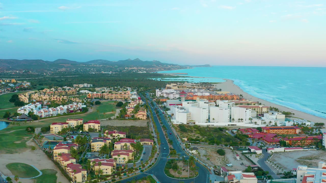 At 0:02, the frame shows San Jose Del Cabo resort buildings and a straight road leading toward the beach, with ocean on the right, mountains on the horizon, and no visible people.