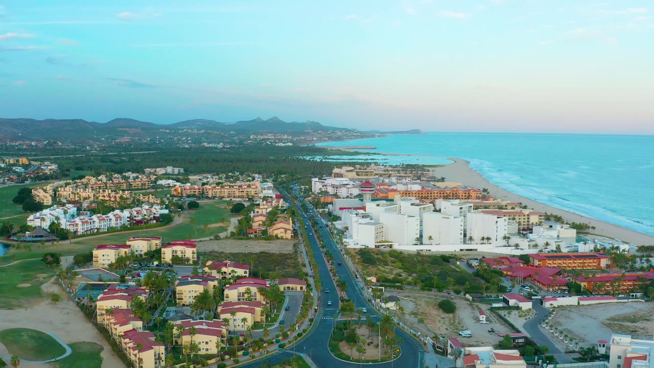 Aerial coastline frame with resort buildings, golf green, road, beach, and blue water.