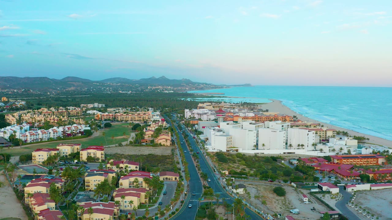 Opening aerial frame over San Jose Del Cabo with a divided road, low residential buildings, white beachside blocks, turquoise ocean, sandy beach, distant hills, and no visible people.