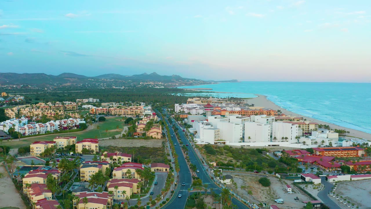 Aerial view in San Jose Del Cabo with the road centered lower in frame, small cars on the lanes, residential roofs in the foreground, ocean to the right, mountains in the distance, and no visible people.