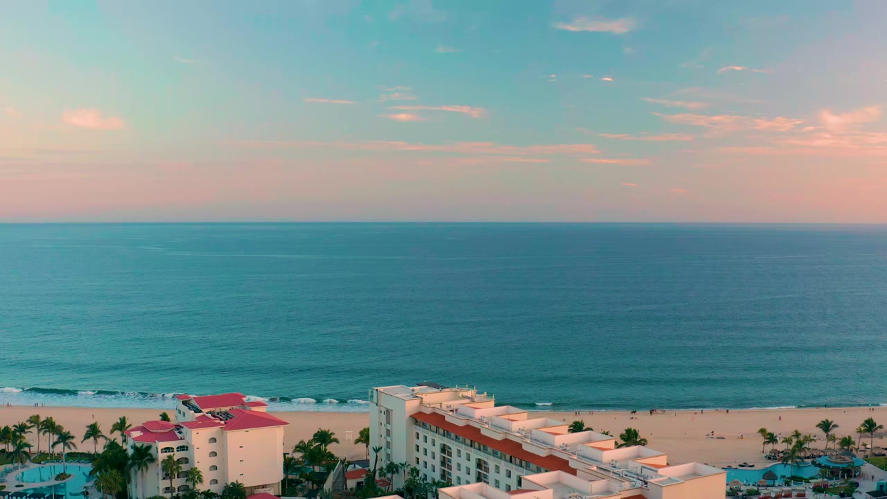San Jose del Cabo beach scene with turquoise ocean, sandy shore, resort roofs, palm trees, pink-blue sky, and no visible people at the start of the clip.