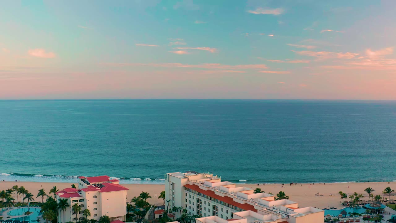 San Jose del Cabo shoreline with the horizon centered high, calm teal water, a narrow sandy beach, resort buildings along the bottom edge, and no visible people.