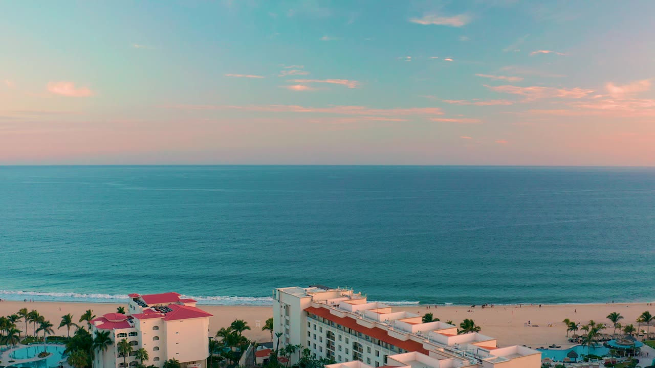 San Jose del Cabo beach and resort view with gentle waves near shore, pale sand, palm-lined buildings at the bottom, pink cloud color, and no visible people.
