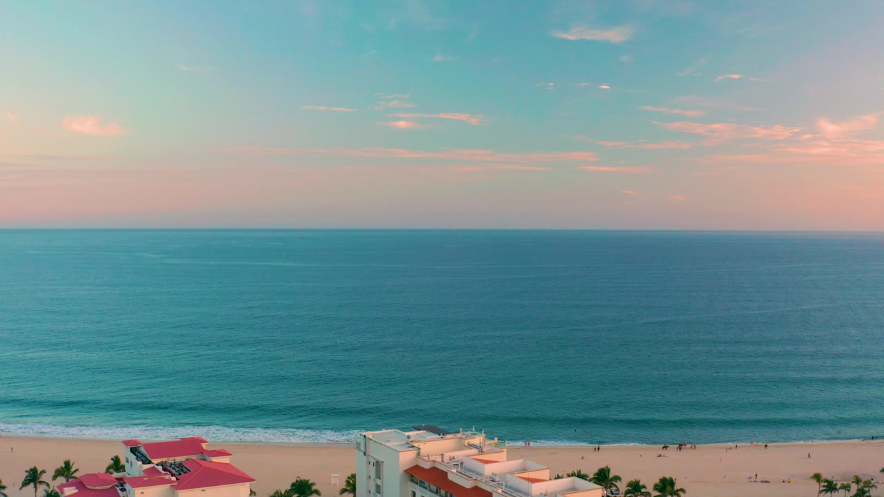 San Jose del Cabo coastline at sunset with turquoise water, sandy beach, palm trees, pale buildings, red roofs, and tiny beachgoers along the shore.