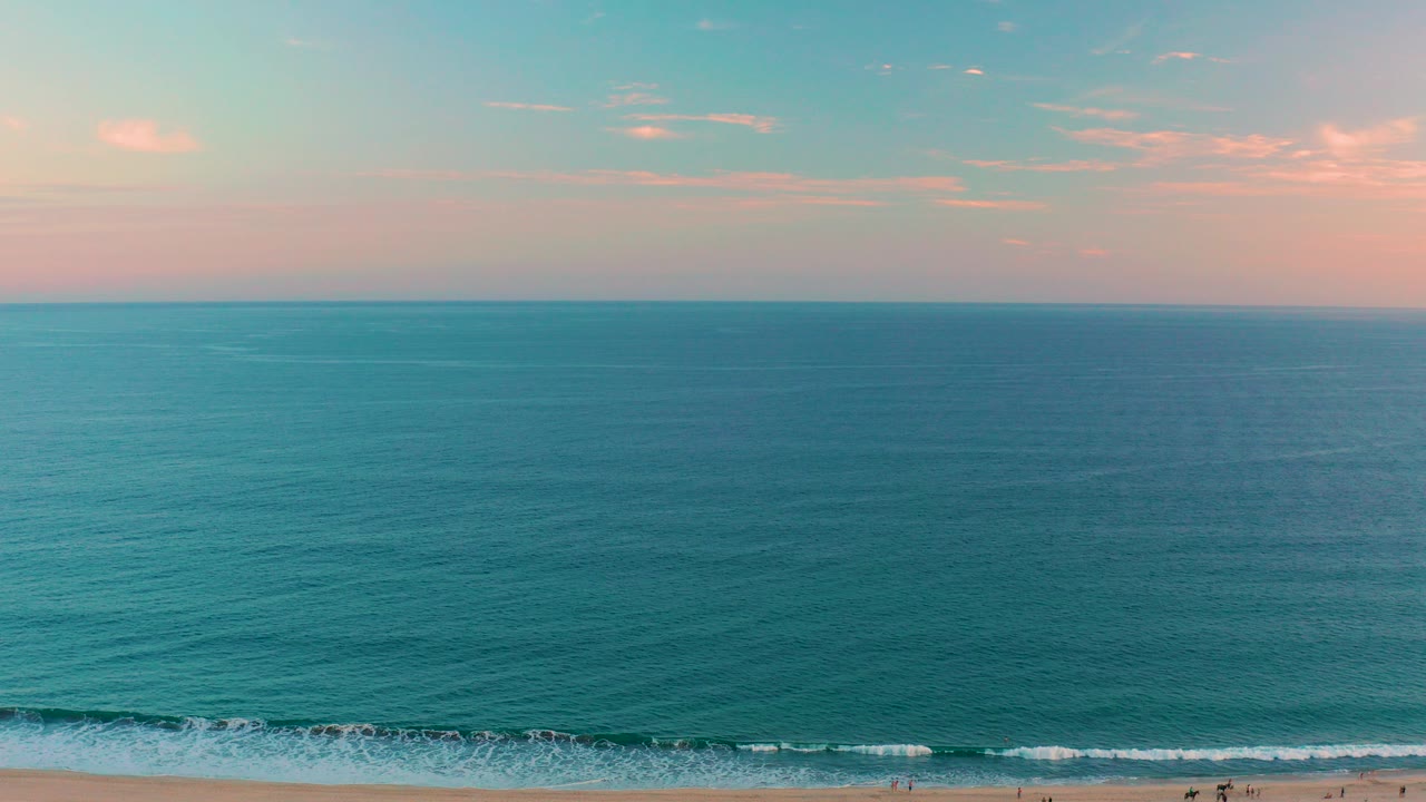 Aerial dusk view over San Jose del Cabo beach, with teal ocean, a narrow surf line, tiny people on the sand, and pink clouds above the horizon.