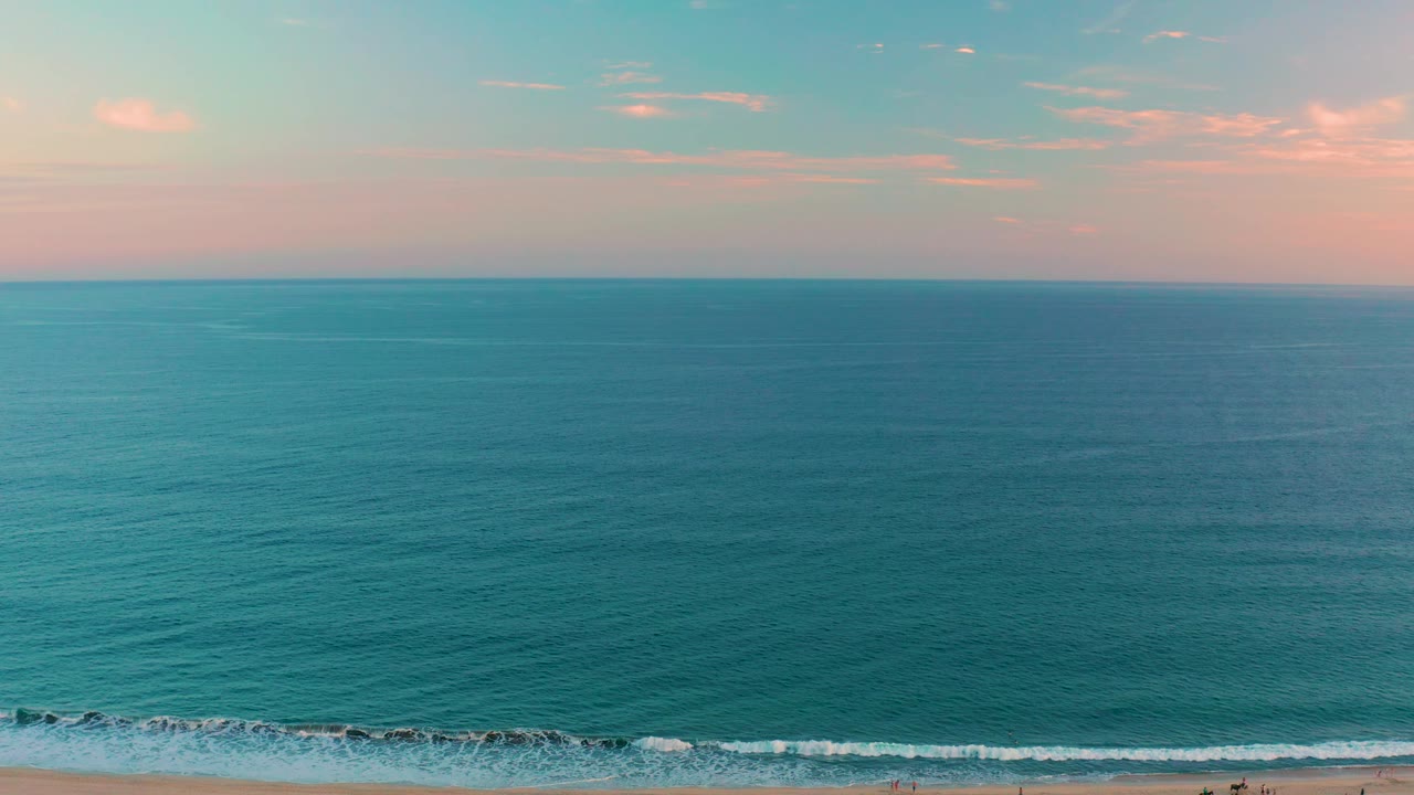 San Jose del Cabo shoreline sits low in frame while teal water dominates, with tiny figures on the sand and a soft blue and pink sky above the ocean horizon.