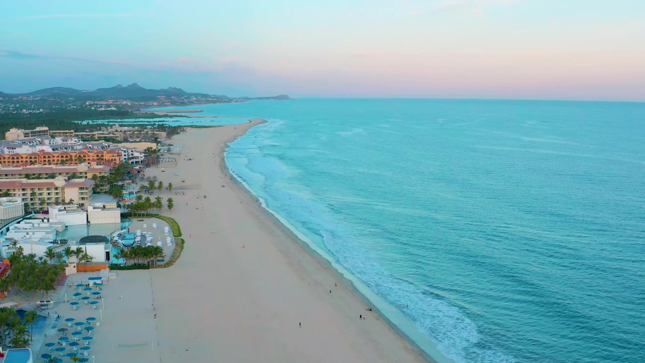 Wide aerial frame in San Jose Del Cabo with gentle surf lines curving onto the sand, coastal buildings grouped at the left edge, open turquoise water to the right, and no visible people.