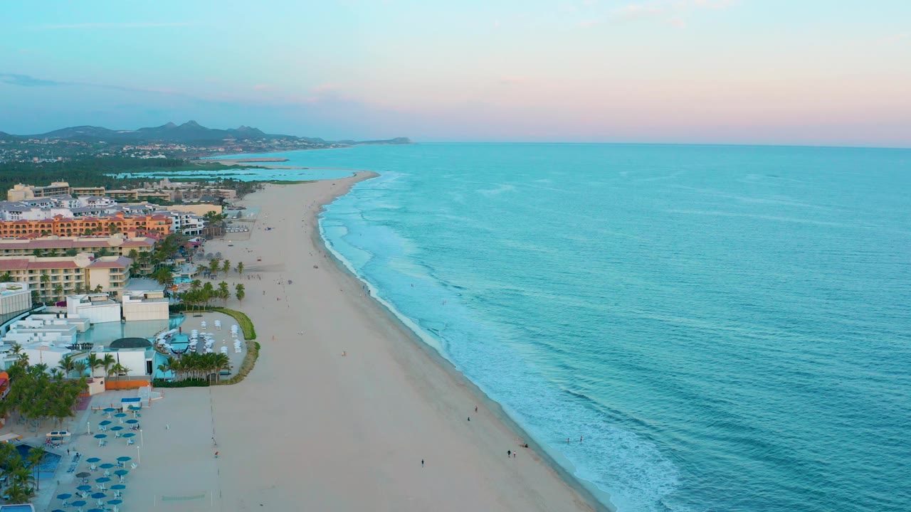 Drone-style aerial over San Jose Del Cabo with the beach running diagonally through frame, blue-green ocean filling the right side, buildings and palms at left, and no visible people.