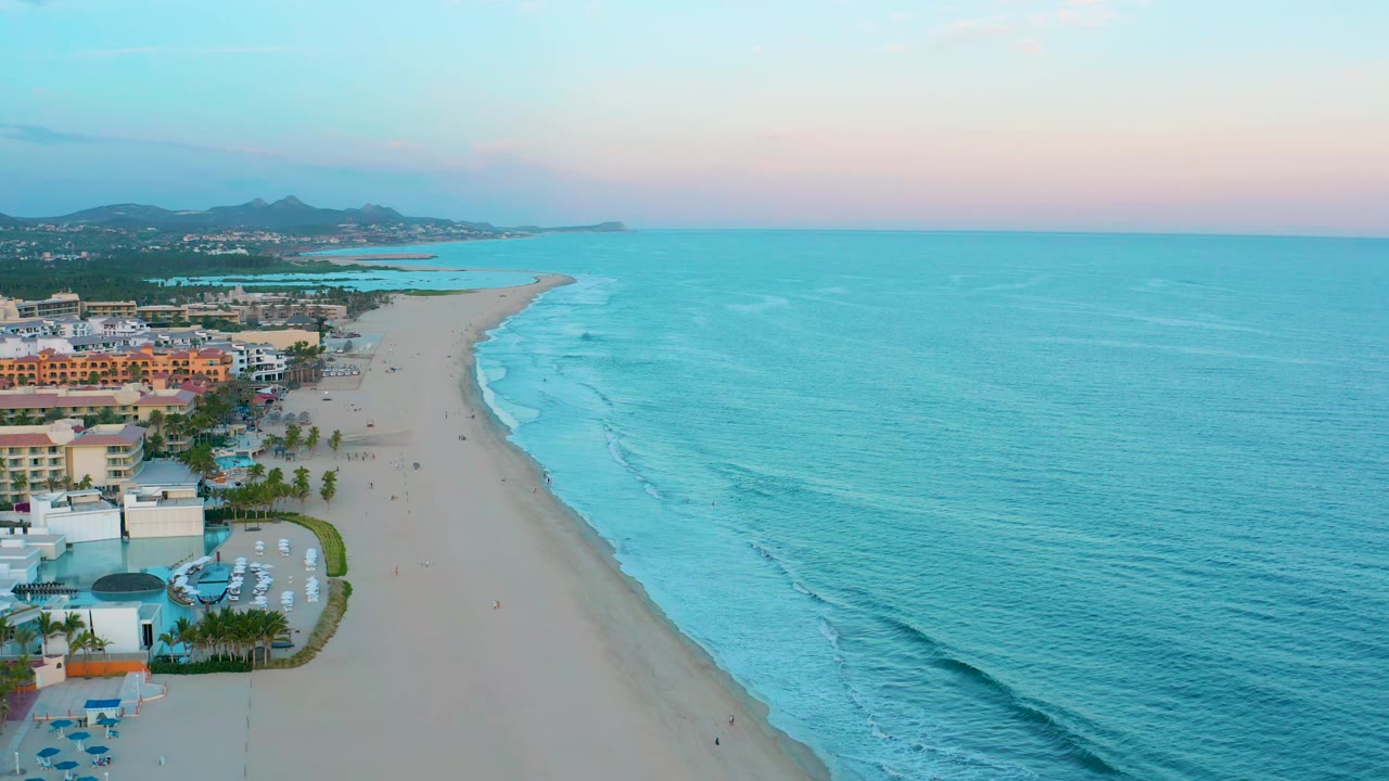 Aerial view over San Jose Del Cabo coastline showing resort pools and palms along the left edge, a long sandy beach, bands of surf, and a pastel evening sky with no visible people.