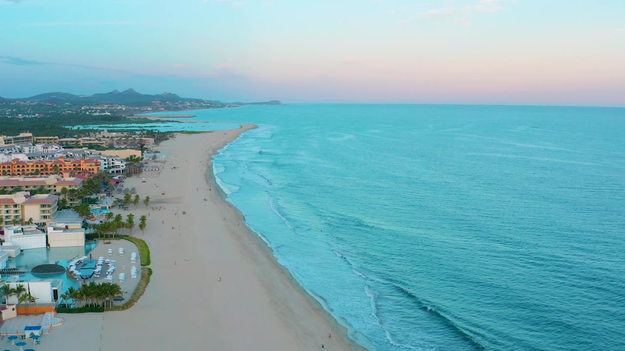 San Jose Del Cabo aerial frame with ocean taking up most of the right side, resort buildings clustered on the left, a curving sandy shoreline, low hills, and no visible people.