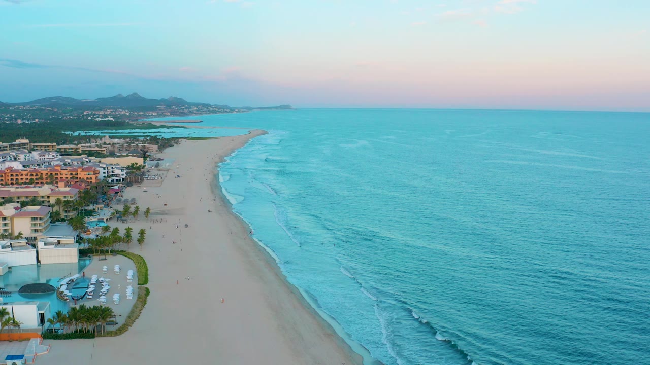 Aerial view along a sandy beach with turquoise waves and resort buildings.