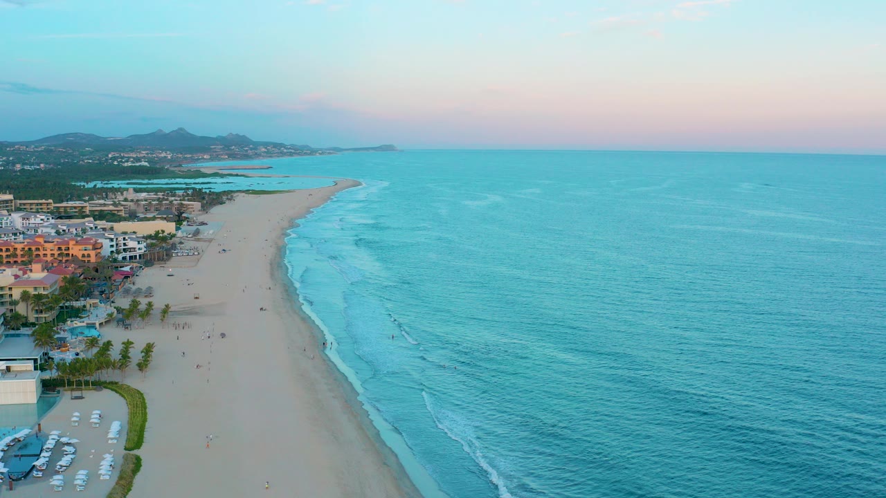 Aerial dusk view in San Jose del Cabo, Mexico, with shoreline buildings and palms at left, a wide sandy beach through the center, and turquoise ocean filling the right side.
