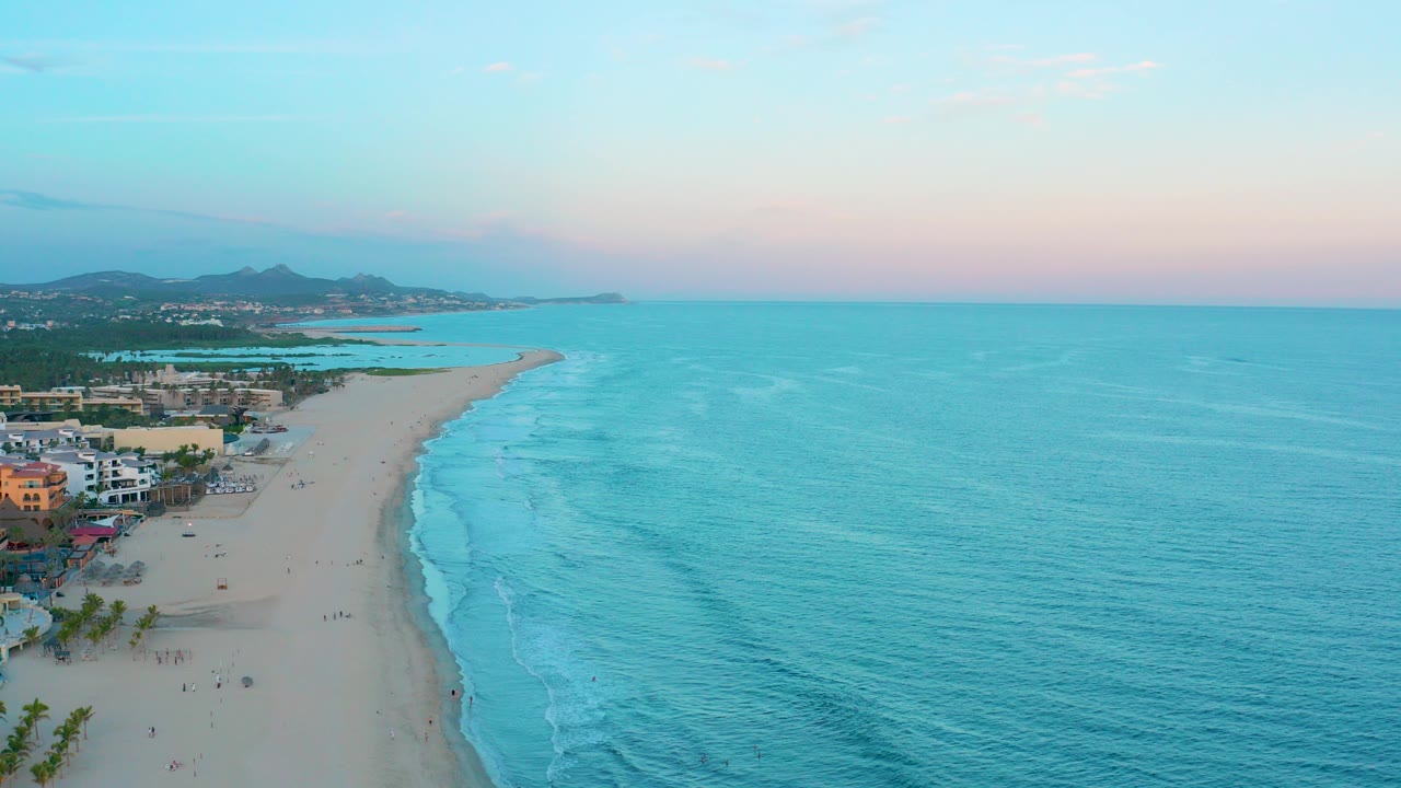Aerial dusk view over San Jose del Cabo, Mexico, with turquoise ocean on the right, a curving sandy beach on the left, shoreline buildings, distant hills, and tiny beachgoers visible.