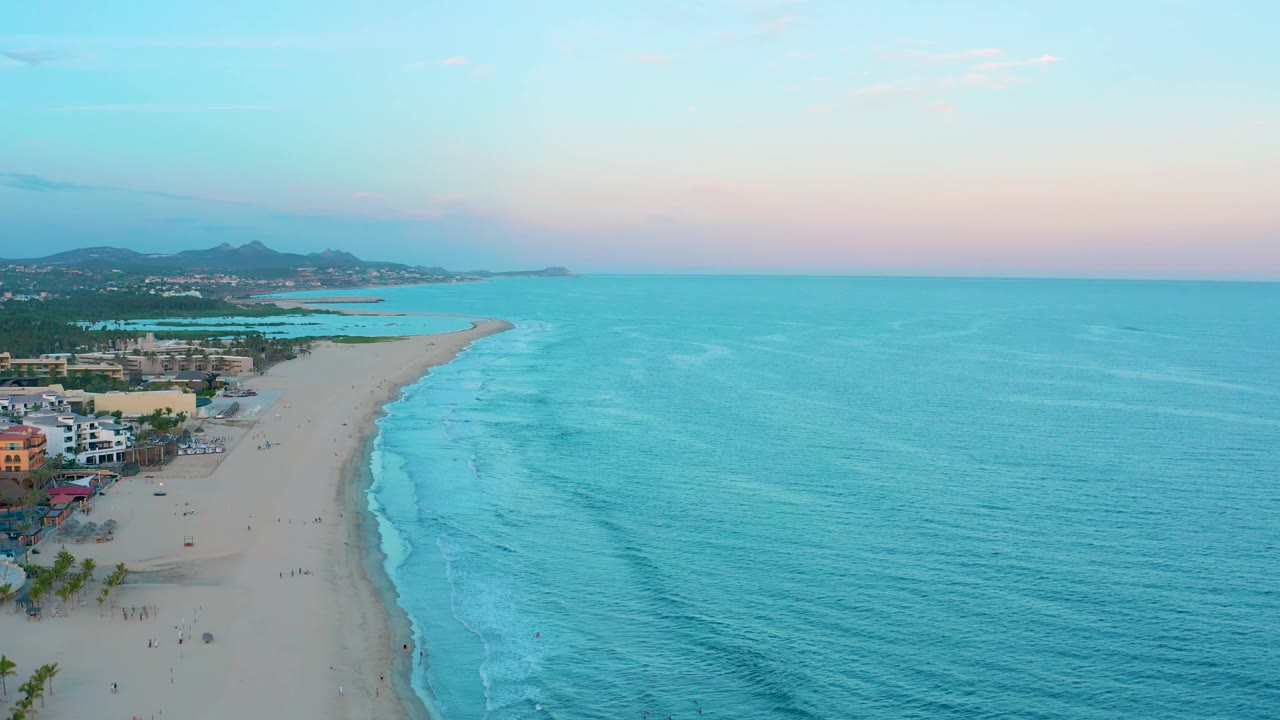 Wide aerial frame of San Jose del Cabo beach at dusk, with open turquoise water dominating the right side, shoreline buildings at lower left, a narrow sand curve, distant hills, and tiny people visible.