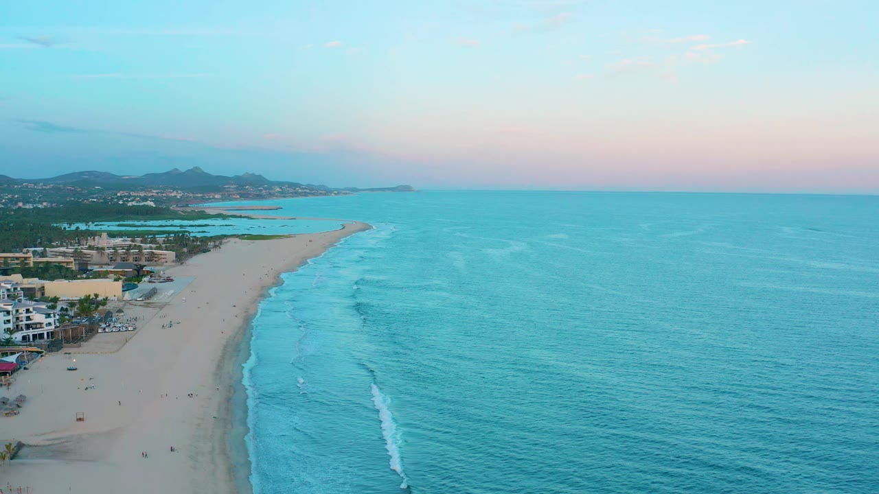 Drone view of a curved beach, pale surf, ocean horizon, and beachfront development.