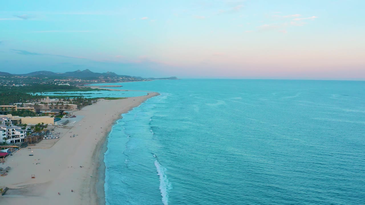 Wide aerial view over the San Jose del Cabo shoreline with open turquoise sea, a narrow line of surf, sandy beach, low buildings at frame left, distant hills, and small figures on the beach.