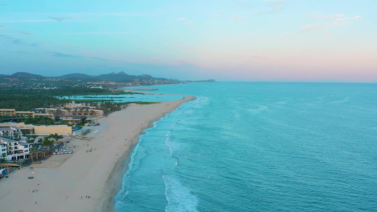 Wide drone view of shoreline with waves, beach buildings, and mountains under a pink-blue sky.