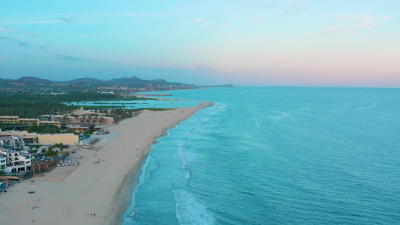 High aerial frame of San Jose Del Cabo beach with turquoise water filling the right side, a broad sand strip through the center, resort-like coastal buildings at left, and small visible people near the waterline.