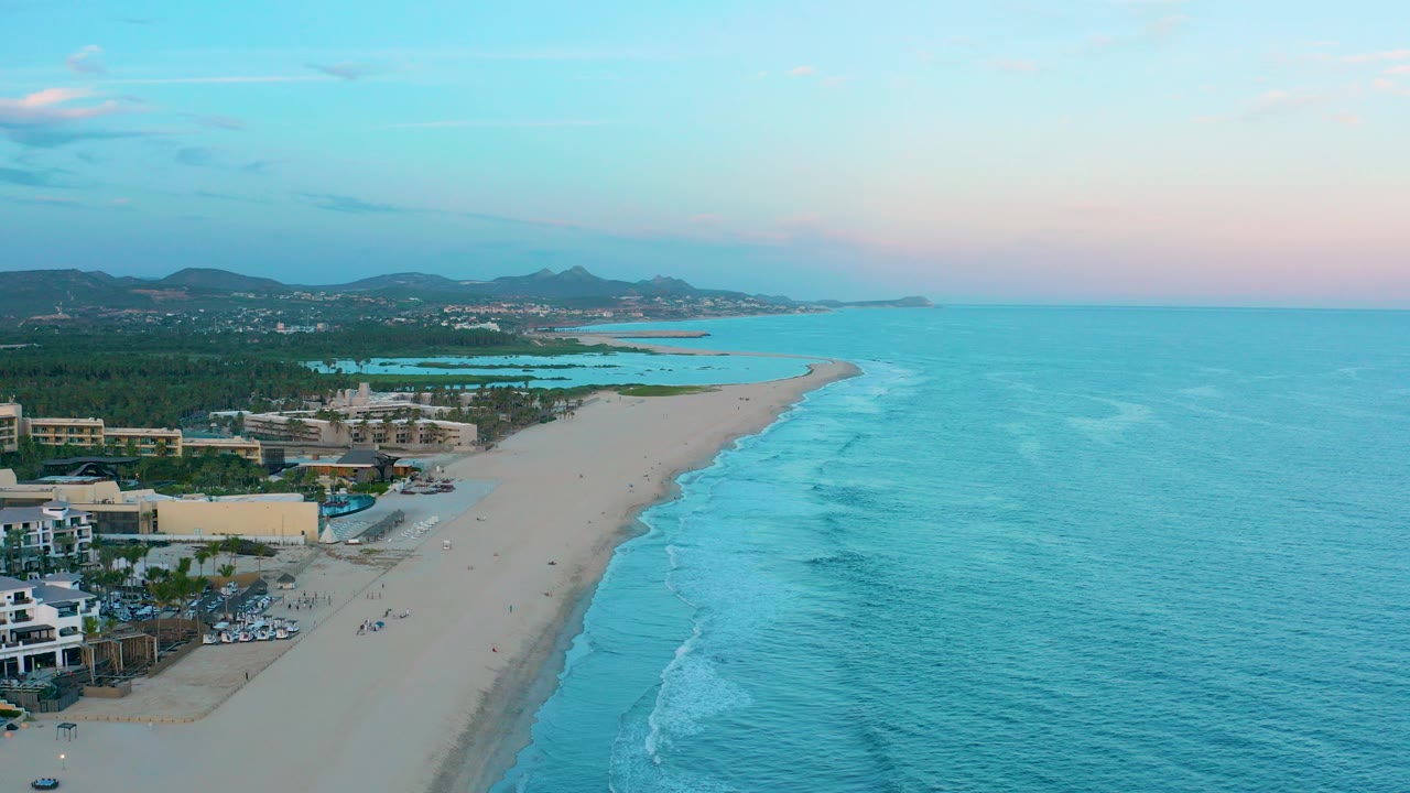 Wide aerial frame over San Jose Del Cabo showing a sandy beach curving along turquoise water, resort buildings at left, low hills in the distance, and tiny people on the sand.